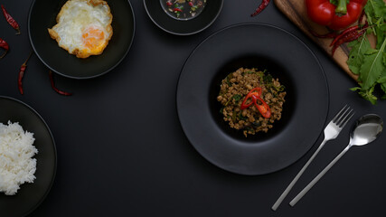 Stir fried minced pork with basil (Pad ka prao), rice fried egg serving on black ceramic plate, silverware and copy space on black table