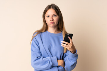 Young Lithuanian woman using mobile phone isolated on beige background with sad expression