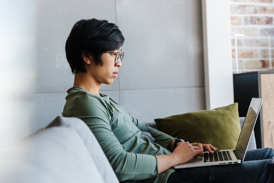 Image of focused asian man working with laptop while sitting