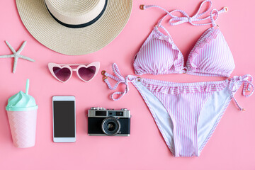 Flat lay of summer items with pastel pink bikini, smartphone, beach hat, camera, pink plastic glass and sunglasses on pink background, top view and copy space. Summer concept.
