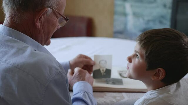 household, kind grandfather with his beloved grandchild shows photos of his youth gladness in photo album of youth during family pastime