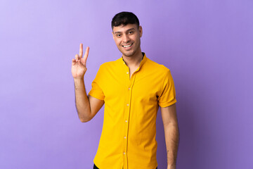 Young Colombian man isolated on purple background smiling and showing victory sign