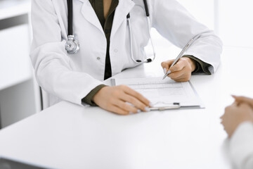 Unknown woman-doctor and female patient sitting and talking at medical examination in clinic, close-up. Therapist wearing green blouse is filling up medication history record. Medicine concept