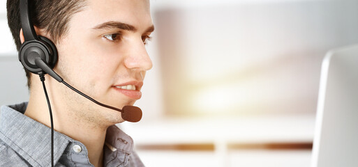 Casual dressed young man using headset and computer while talking with customers online in sunny office. Call center, business concept