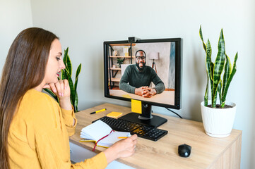 Video meeting with with an african-american coworker, employee. A young woman sits at office desk and talking with young man via video call, she takes a notes