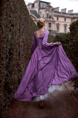 Fototapeta premium A beautiful blonde girl model runs in a purple long flowing dress in a maze park in Paris, surrounded by green bushes