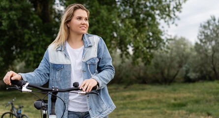 Fototapeta na wymiar bike ride in the park. Young beautiful woman is standing in the park holding a bicycle in her hands. Denim clothing. weekend break.