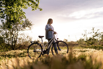 Obraz premium A young woman is walking near a bicycle in a city park at summer sunset. Blurred background