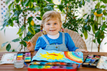 little toddler girl painting rainbow and sun with water colors during pandemic coronavirus quarantine disease. Children painting rainbows around the world with the words Let's all be well. Happy child