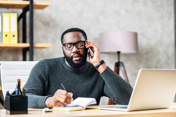 Young black man is solving business tasks on the phone, concentrated guy sits at the desk with...