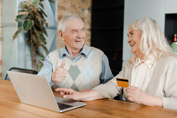 cheerful elderly couple showing thumb up while shopping online with credit card and laptop at home during self isolation