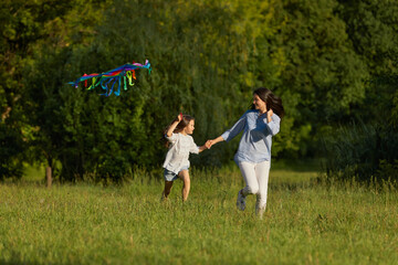 Naklejka na ściany i meble smiling mom and child daughter run with kite in park in sunny summer day. Carefree time