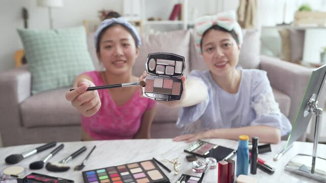 two makeup artist bloggers demonstrating cosmetic product live online streaming face camera. charming asian chinese women friends smiling recording video of beauty channel and showing pink palette