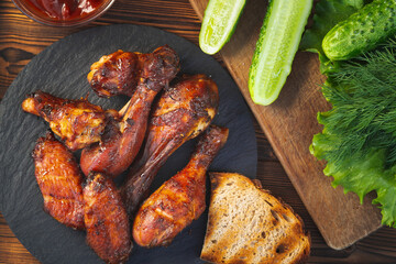 Grilled chicken drumsticks and wings on a black slate dish with fresh cucumbers, tomato sauce and herbs, top view, flat lay