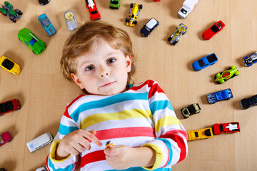 Lovely blond kid boy playing with lots of toy cars indoor. Happy healthy child boy having fun during pandemic coronavirus quarantine disease. Child alone at home, closed nursery.