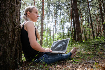 girl in the forest sits leaning against a tree, working on a laptop. rest at nature. in the background blurred silhouettes of trees. digital detox. Mental health