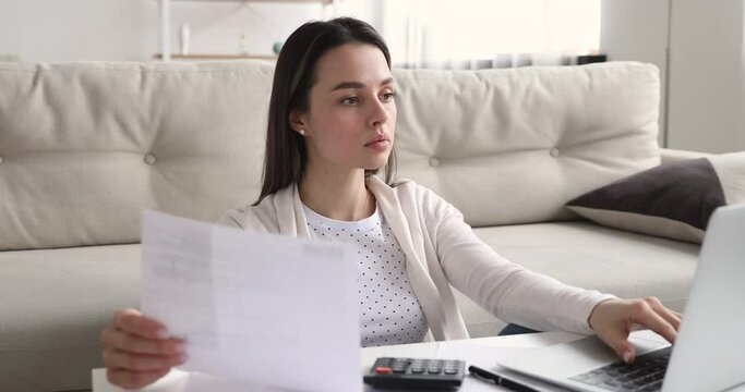 Concentrated young woman holding paper receipt, checking taxes information using e-banking application on computer. Smart millennial lady managing monthly budget, rental payments or utilities bills.