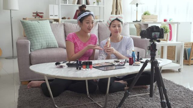 two Asian female bloggers recording vlog video with makeup cosmetic at home living room. girl friends online influencer on social media concept. women talking on live streaming to camera on tripod