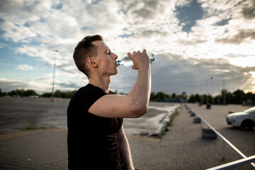 Young man standing on sports ground during morning workout and drinking water from a bottle.