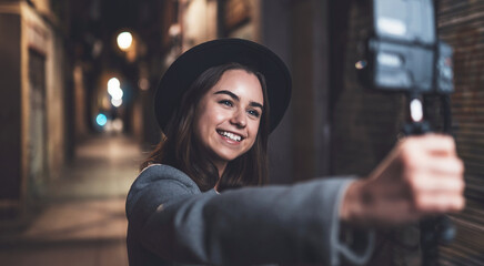 Traveler female blogger shooting video for social media with digital camera. Smiling young woman vlogger taking selfie video on light night city