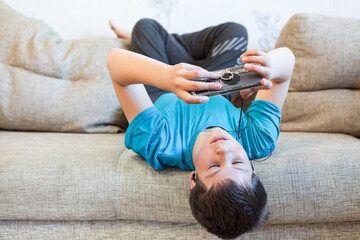 Portrait of Caucasian teenager with large smartphone or game console in hands, playing or engaging over social media while lying his back on sofa