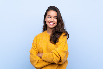 Young Colombian girl with sweater over isolated blue background with arms crossed and happy