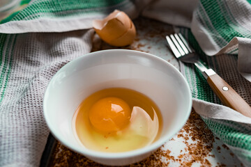 Raw egg broken into a white bowl with yolk and protein, next to the shell, fork and towel on a rusty metal background as at home in the village, everything simple and ordinary 