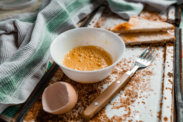 Whipped with a fork and mixed raw egg broken into a white bowl with yolk and protein, next to the shell, fork and towel on a rusty metal background as at home in the village, everything simple and ord