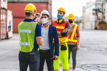 Professional of group cargo foreman in helmets standing and using infrared thermometer for checking body temperature staff fever before work in quarantine for coronavirus wearing protective mask