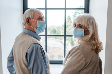 Obraz premium sad senior couple in medical masks standing near window during self isolation