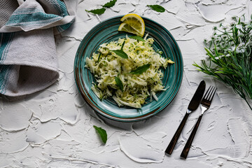Crispy salad of fresh vegetables with herbs. Young cabbage with cucumber and arugula leaves, decorated with lemon and a bouquet of white flowers on a light textured stone table with Cutlery
