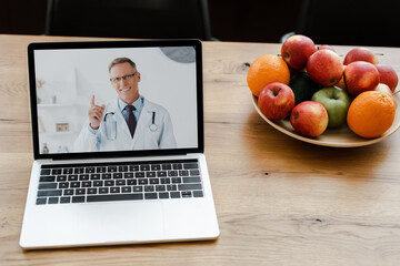 close up of laptop with smiling doctor on screen on table with fruits