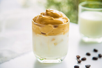 Close up a cup of Dalgona coffee with blurred coffee beans and a cup of milk on a white table with a natural blurred background in the afternoon.