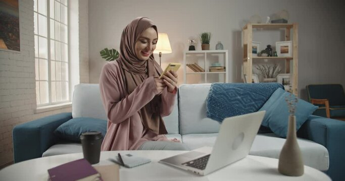 Young pretty girl wearing traditional hijab head scarf is using her smartphone, scrolling through social media and positively smiling - modern muslims, communication concept 4k footage