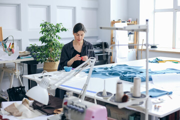 Caucasian young woman fashion designer sewing, stitching blue fabric, using needle string. Interior of light workshop studio.