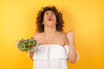 Portrait of beautiful blonde female looks with excitement up, keeps hands raised, notices something unexpected, poses indoors. Lovely woman reacts on sudden news.