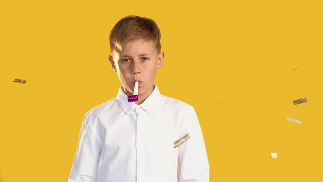 Sad boy portrait. Birthday alone. Disappointed kid blowing horn in confetti rain isolated on orange background.