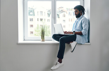 Thoughtful young African man using laptop while sitting on the window sill indoors