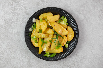 Roasted, baked potatoes on a black plate with fresh green onions and rosemary. Light gray background. Top view. Copy space.