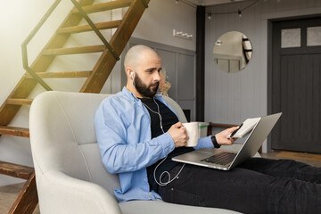 A guy in a blue shirt and black jeans is sitting on the couch and talking on a video link at a computer and drinking a cup of coffee. The concept of freelance work