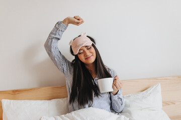 Long-haired woman in blue pajamas streches sweetly after sleeping. Girl holding cup