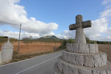 A summer day shot of an old stone cross on the side of a country road with a field with olive trees, a mountain and a blue sky with white clouds in the background. Mallorca island, Spain