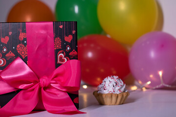 Fototapeta na wymiar festive cupcake on the background of red gift box with a pink ribbon, multi-colored balloons and lights. party, birthday concept. Copy space. . High quality photo