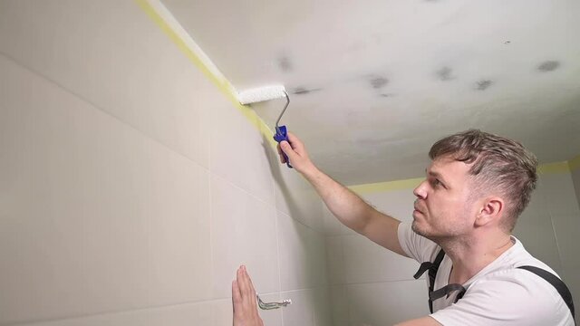 Worker painting ceiling with white paint. Painter man paints bathroom ceiling with roller tool. Handyman type using paint roller. Repair in apartment. Homeowner making renovation repair at home.