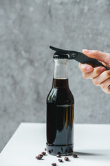cropped view of woman opening cold brew coffee in bottle with opener on white table with coffee...