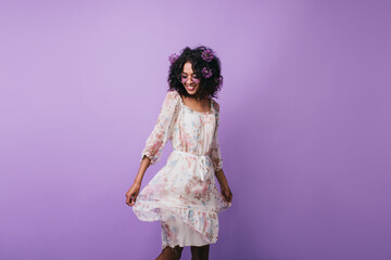 Amazing dark-haired girl posing in trendy summer dress in studio. Portrait of gorgeous african woman dancing with inspired smile on purple background.