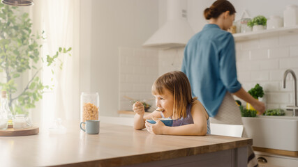 Obraz na płótnie Canvas Breakfast in the Kitchen: Adorable Little Daughter Eating with Pleasure, Young Beautiful Mother Cooking in the Background. Caring Mother Prepares Cereal Breakfast for Her Cute Little Girl