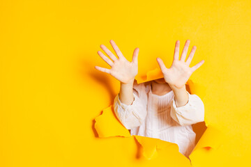 Child hands is showing ten fingers through a ripped hole in yellow paper, with copy space.
