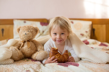 Cute blonde toddler child,reading book and eating croissant in bed, laughing