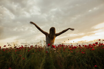 Girl in poppy field with hands up on sunset. Happy girl with raised up hands standing  and looking up in the sky, enjoying summer vacation. Free happy woman raising arms watching the sunset.Copy space